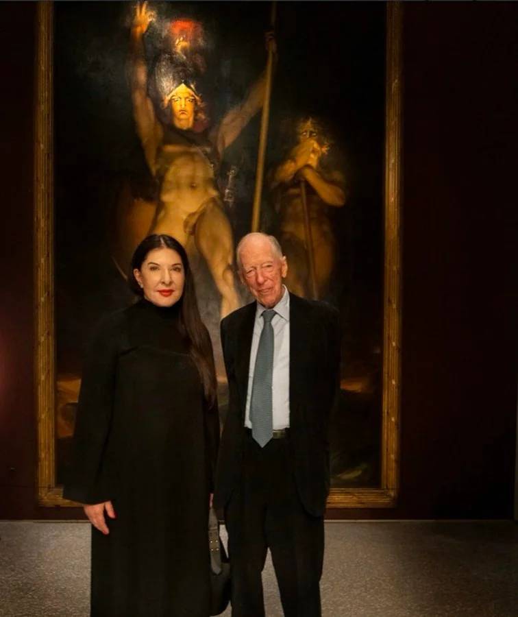 Marina Abramović y Jacob Rothschild, de pie y vestidos de negro, ante el cuadro Satanás convocando a sus legiones, de Sir Thomas Lawrence, en el que Satanás aparece desnudo, con el brazo derecho en alto y una lanza en el izquierdo.
